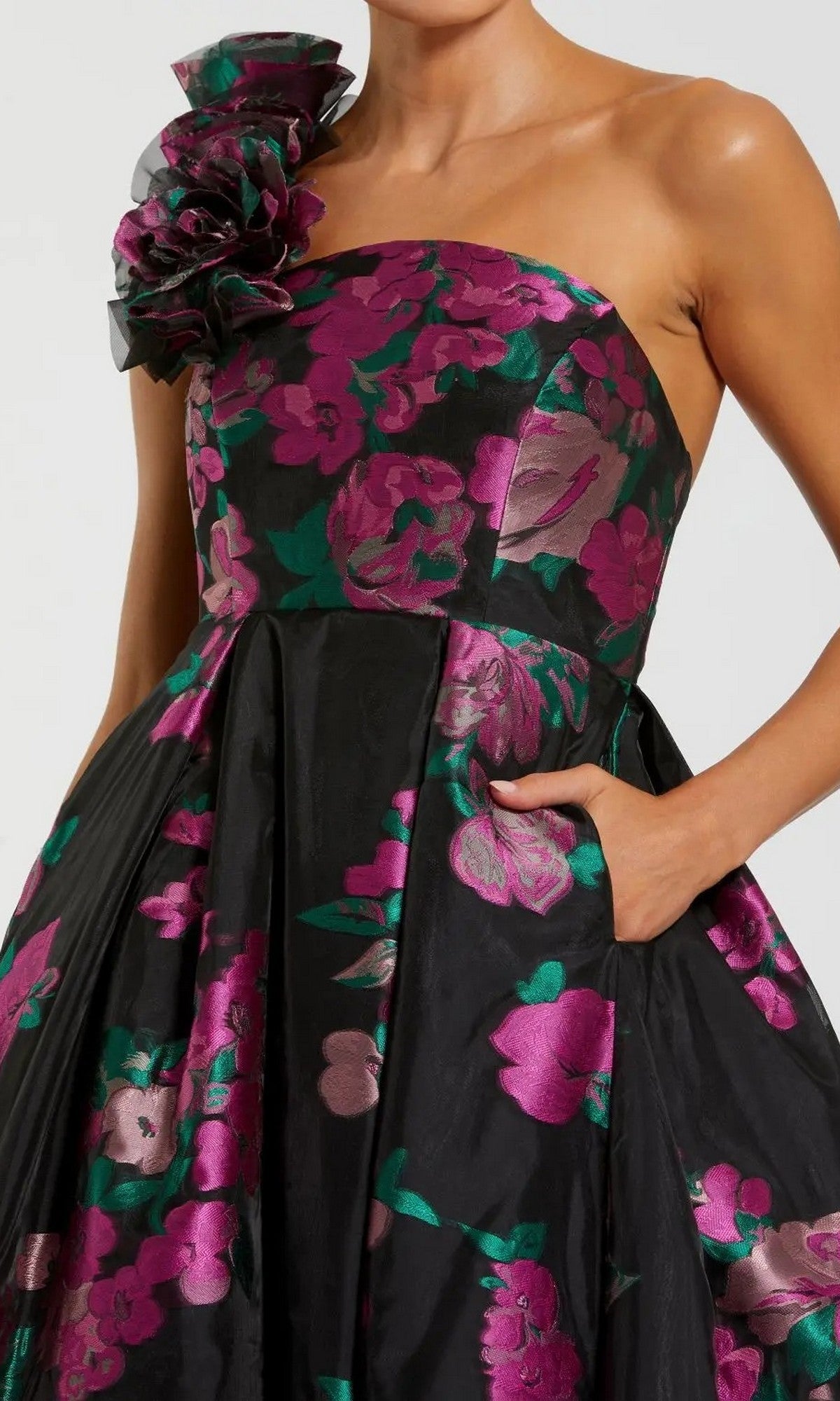 A woman wears the Mac Duggal 11967 long formal dress, a strapless black ballgown featuring pink and purple floral patterns and a large fabric flower detail on one shoulder.